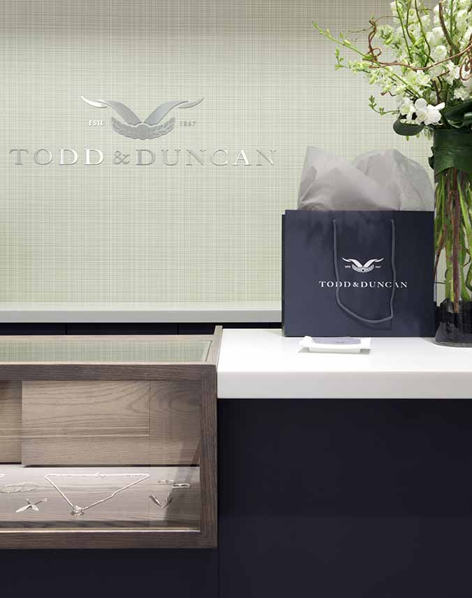 A blue Todd & Duncan gift bag sitting on a counter next to a vase of flowers, with the Todd & Duncan name and logo in metal on the wall behind the counter.